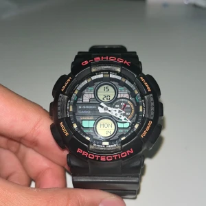Svart Casio G-Shock GA-140 klocka - Säljer en robust och sportig svart Casio G-Shock GA-140 med digital och analog display. Klockan har röd och orange text på boetten, flera funktioner som tidtagning och datum, samt tåligt armband i syntetmaterial. Perfekt för dig som gillar en tuff och teknisk stil. (Helt ny batteri)