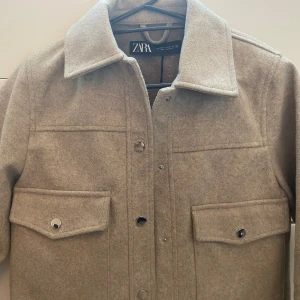 Beige kappa - Snygg beige ”kappa” från Zara i ullmix med klassisk krage, stora bröstfickor med lock och silvriga tryckknappar. rak passform och lång ärm. Enkel att styla till både jeans och kjol.
