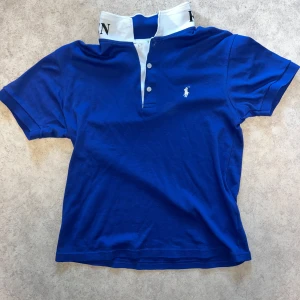 Blå Polo Ralph Lauren t-shirt - Snygg blå t-shirt från Polo Ralph Lauren med vit krage och knappar framtill. Klassisk broderad logga på bröstet och stor text på insidan av kragen. Skön passform och tillverkad i mjuk bomull. Perfekt för en stilren och sportig look.