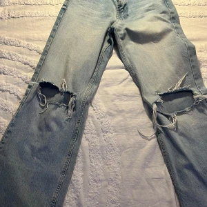 Ljusblå ripped jeans från Zara - Säljer ett par ljusblå jeans från Zara med stora slitningar och hål över knäna. Jeansen har rak passform och klassisk femficksdesign. Perfekta för dig som gillar en edgy och avslappnad stil.