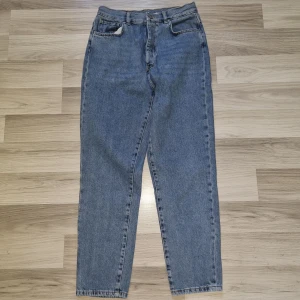 Never Denim blå jeans, storlek M - Klassiska Never Denim jeans i mellanblå tvätt med raka ben och hög midja. Jeansen har knäppning med knappar, fem fickor och är tillverkade i robust bomullsdenim. Perfekta för en avslappnad och tidlös stil.