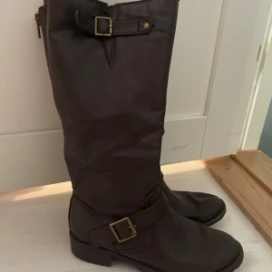 Bruna höga boots med spännen och dragkedja - Snygga bruna boots i skinn med högt skaft, dekorativa spännen i mässing och röda dragkedjor baktill. Platt sula och rund tå ger en klassisk look. Perfekta för höst och vinter när du vill ha en cool och tidlös stil.