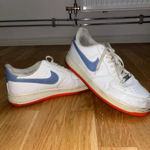 Nike Air Force 1 med blå swoosh - Snygga Nike Air Force 1 sneakers i vitt skinn med ljusblå swoosh och orange sula. Klassisk låg modell med snörning och perforerad tå för extra ventilation. Perfekta för dig som gillar retrovibbar och streetstyle. (Ej original)