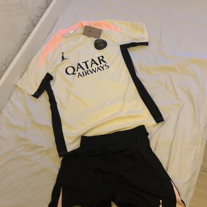 PSG x Jordan fotbollströja beige/svart - Snygg PSG fotbollströja från Jordan i beige med svarta detaljer och ljusrosa axelpartier. Tröjan har korta ärmar, rund hals och är tillverkad i lätt, ventilerande material. Stort Qatar Airways-tryck på bröstet och PSG-logga. Perfekt för dig som älskar fotboll och streetstyle.