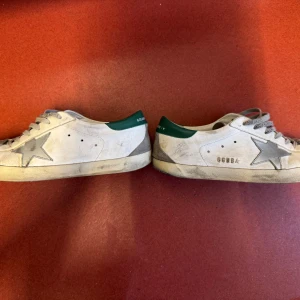 Golden Goose Superstar sneakers vit/grå - Golden Goose Superstar sneakers i vitt skinn med grå detaljer och grön häl. Klassisk stjärndesign på sidan, grå snörning och svart sula. Snyggt slitna för en cool, avslappnad look. Perfekta för dig som gillar streetstyle och unika detaljer.