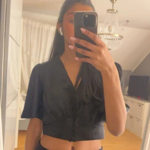 Svart croppad blus från H&M - Snygg svart croppad blus från H&M i glansigt material med v-ringning och klädda knappar framtill. Endast testad! 