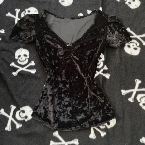 Svart handsydd sammetsblus med ros - Supersöt svart sammetsblus med korta puffärmar och v-ringning. Framtill sitter en liten svart rosett med blomdetalj. Blusen har figurnära passform och mjuk, glansig yta som ger en lyxig känsla. Perfekt för dig som gillar en lite goth eller romantisk stil. Plagget är handsytt, vilket innebär att det kan förekomma mindre skavanker tex lösa trådar! 🪡 