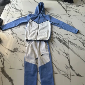 Nike tech - Nike träningsdress i storlek L med hoodie och dragkedja. Dressen är tvåfärgad i ljusblått och vitt med svarta detaljer och Nike-logga på både jacka och byxa. Mjukt material och bekväm passform, perfekt för sport eller chill.