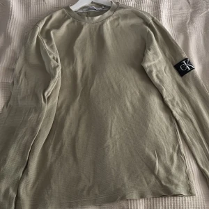 Beige långärmad tröja Calvin Klein - Snygg beige långärmad tröja från Calvin Klein Jeans med diskret struktur i tyget och CK-logga på ärmen. Tröjan har rund halsringning och är perfekt för en clean och stilren look. Passar dig som gillar enkel men trendig stil.