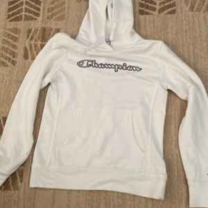 Vit hoodie från Champion med logga - Vit hoodie från Champion med stor logga över bröstet och klassisk känguruficka. Tröjan har huva med snörning och lång ärm, samt broderad Champion-logga på ärmslutet. I storlek M men skulle säga att de är mer för personer som har S/xs
