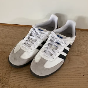 Adidas Samba vita och svarta sneakers - Klassiska Adidas Samba sneakers i vitt och svart med de ikoniska tre ränderna på sidan. Ovandel i skinn och mocka, svart sula och snörning framtill. Använts lite. Storlek 46. Skriv gärna om det är något du undrar!