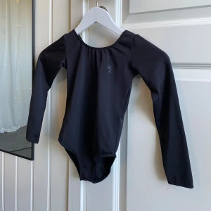 Svart gymnastikdräkt  - En svart långärmad body med rundad halsringning, perfekt för dans eller gymnastik. Tillverkad i stretchigt material som ger bra rörelsefrihet. Enkel och stilren design utan synliga detaljer förutom en liten logga på bröstet.