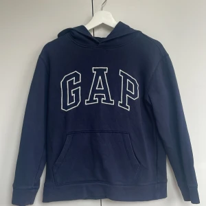 Mörkblå hoodie från GAP Kids - Mörkblå hoodie från GAP Kids med stor vit logga framtill. Tröjan har huva och en klassisk känguruficka. Perfekt för en avslappnad och sportig stil. Liten i storleken och lite korta armar.