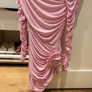 Ljusrosa draperad klänning med blomdetalj från H&M - Supersnygg ljusrosa kortklänning från H&M med draperad design och lång ärm. Klänningen har en markerad blomdetalj i midjan och figurnära passform. Perfekt för dig som vill sticka ut med en trendig look!