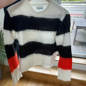 Randig stickad tröja i svart, vitt och rött/orange - Säljer en stickad tröja med breda ränder i svart, vitt och rött. Tröjan har lång ärm och en croppad passform med rå kant nertill. Perfekt för dig som gillar färg och mönster! Bra skick dock lite nopprig men går nog att ta bort med en noppa maskin! Köpt på Vero Moda, märke är Twintip!
