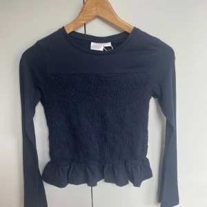 Mörkblå långärmad topp med smock och volang - Säljer en mörkblå långärmad topp med smockad front och rygg samt volangkant nedtill. Perfekt för dig som gillar en söt och trendig stil. Mjuk och stretchig, passar till mycket! Helt ny med prislapp kvar