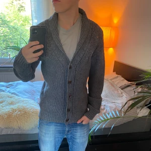 Grå stickad kofta med sjalkrage - Säljer en sjukt snygg grå stickad kofta med sjalkrage och snyggt mönster upptill. Koftan har knappar framtill och ribbade muddar vid ärmslut och nederkant. Perfekt för lager-på-lager, chill vardagsstil och sommar kvällar. Koftan har inga större tecken på användning och befinner sig därav i skick 9.5/10. Tveka inte på att höra av dig vid frågor eller funderingar! ⭐️