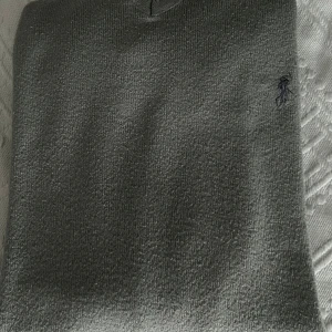 Grå v-ringad stickad tröja från Ralph Lauren - Säljer en klassisk grå stickad tröja från Ralph Lauren med v-ringning och diskret broderad logga på bröstet. Tröjan har långa ärmar och är perfekt för lager-på-lager-stil.