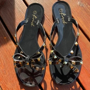 Svarta sandaler med rosett och nitar  - Säljer ett par svarta sandaler från Krush med dekorativa rosetter och guldfärgade nitar. Skorna har öppen tå och platt sula, perfekta för varma dagar. Sandalerna/flippfloppen är aldrig använda och är därför i ett mycket bra skick! 