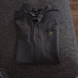 Mörkgrå pikétröja från Lacoste - Säljer en klassisk mörkgrå pikétröja från Lacoste med den ikoniska krokodilloggan på bröstfickan. Tröjan har krage och knäppning framtill. Perfekt för en stilren och avslappnad look.