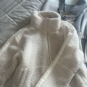 Vit teddyjacka från Kamsic - Mysig vit teddyjacka med hög krage och dragkedja framtill. Jackan har en fluffig och mjuk yta som ger en trendig look. Perfekt för kyliga dagar när du vill vara både varm och snygg. Bra skick förutom att det har blivit fläckar på insidan av jackan, men inget som syns när man har på sig den!