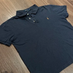 Mörkblå pikétröja från Polo Ralph Lauren - Säljer en klassisk mörkblå pikétröja från Polo Ralph Lauren med broderad logga på bröstet. Tröjan har korta ärmar, krage och knappar framtill. Slim fit-modell i mjuk bomull, perfekt för en stilren look. Står storlek M men passar som XXS