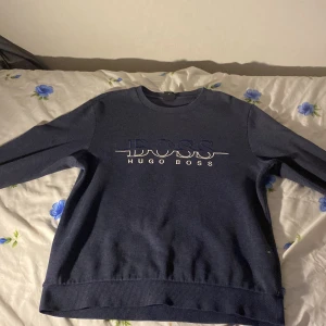 Mörkblå sweatshirt från Hugo Boss - Säljer en mörkblå sweatshirt från Hugo Boss med vit logotyp framtill. Tröjan har rund halsringning och långa ärmar. Klassisk och stilren design, perfekt för dig som gillar märkeskläder.