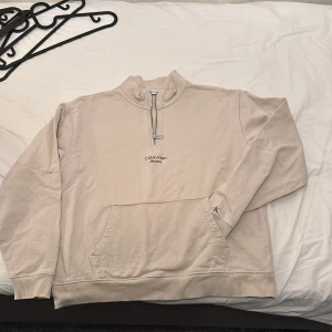 Beige halvzip sweatshirt från Calvin Klein Jeans - Säljer en beige sweatshirt från Calvin Klein Jeans med halv dragkedja och broderad logga på bröstet. Tröjan har en stor magficka och ribbade muddar vid ärmslut och nederkant. Perfekt för en avslappnad stil.