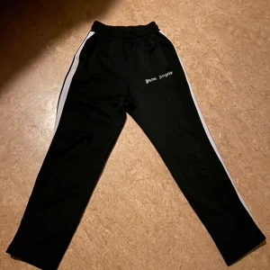 Svarta mjukisbyxor från Palm Angels med vita revärer - Svarta mjukisbyxor från Palm Angels med vita revärer längs sidorna och logga på vänster lår. Byxorna har elastisk midja och raka ben, perfekta för en avslappnad stil.