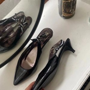 Kitten heels - Säljer ett par svarta kittenheels i lackat material med spetsig tå. Skorna har en söt rosettdetalj på sidan och öppen häl med rem. Skitsnygga, men tyvärr aldrig använda🫦