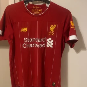 Liverpool FC hemmatröja - Liverpool FC:s klassiska röda hemmatröja från New Balance med vita tunna ränder, klubbmärke och sponsortryck på bröstet. Kortärmad modell med vita detaljer vid ärmslut och krage. Perfekt för alla fans! Skriv för funderingar eller frågor💯💯💯