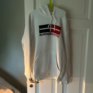 Vit hoodie från H&M med tryck - Vit hoodie från H&M med trycket 'SURFACE EARTH' och en grafisk flagga på bröstet. Tröjan har huva med dragsko och en stor magficka. Perfekt för en avslappnad stil.
