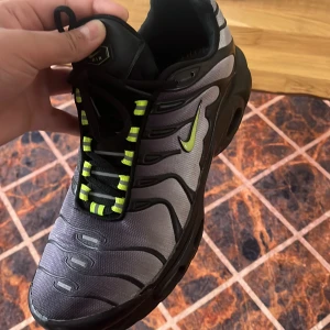 Nike sneakers med neongula detaljer - Säljer ett par Nike sneakers i svart och grått med neongula detaljer på snörningen och Nike-loggan. Skorna har en sportig design med rund tå och platt sula, perfekta för en streetwear-look.