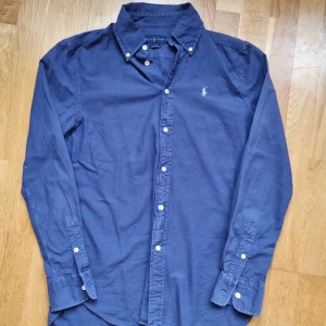 Blå skjorta från Ralph Lauren - Klassisk blå skjorta från Ralph Lauren med vit broderad logga på bröstet. Skjortan har långa ärmar, knappar framtill och vid ärmslut. Perfekt för en stilren och tidlös look.
