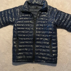 Peak performance jacka  - Säljer en mörkblå dunjacka från Peak Performance med dragkedja framtill och två sidofickor med dragkedja. Jackan är lätt och smidig, perfekt för lite kallare dagar och kyliga sommarkvällar. Klassisk design med quiltat mönster och hög krage. Passar 158-165