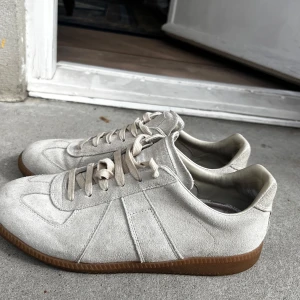 Maison Margiela sneakers i mocka med gummisula - Storlek 43 men passar även 44. Säljer ett par stilrena beige sneakers i mocka med klassisk design och bruna gummisulor. Skorna har snörning och rund tå, perfekta för en clean och avslappnad look. Passar till det mesta och är riktigt bekväma. Toppen skor för våren sommaren. Skorna är lite smutsiga, men det går enkelt att få bort med en mocka borste.