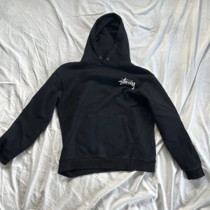 Svart hoodie från Stüssy med tärningsmotiv Medium  - Säljer en svart hoodie från Stüssy med vit logga på bröstet och stort tryck med tärningar på ryggen. Klassisk känguruficka och huva med dragsko. Perfekt för dig som gillar streetwear,Grisch och vill sticka ut med snygga detaljer.