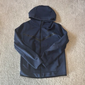 Mörkblå Nike tech  hoodie Nike - Säljer en mörkblå hoodiejacka från Nike med dragkedja och huva. Hoodie har diskreta reflexdetaljer på framsidan och Nike-logga på bröstet. Tillverkad i mjukt syntetmaterial, perfekt för träning eller vardag.