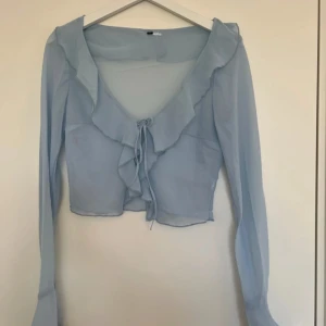 Ljusblå volangblus med knyt - Supersöt ljusblå blus med volangkant och knyt framtill. Blusen är genomskinlig och har långa ärmar, perfekt för en trendig och feminin look.