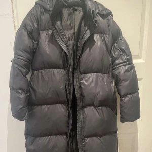 Svart lång pufferjacka med huva - Säljer en svart lång pufferjacka med huva och bälte i midjan. Jackan har dragkedja framtill och är perfekt för kalla dagar. Den är vadderad och har en oversized passform som ger en cool look.