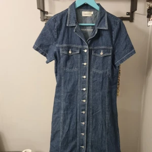 Jeansklänning med knappar från Cappuccini - Snygg jeansklänning från Cappuccini med korta ärmar och klassisk krage. Klänningen har knappar framtill och två bröstfickor med lock. Perfekt för en avslappnad och trendig look.