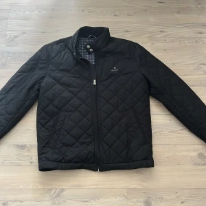 Svart quiltad jacka från Gant - Stilren svart quiltad jacka från Gant med dragkedja framtill och ståkrage med knappdetalj. Nypris på dessa ligger på runt 2200kr 