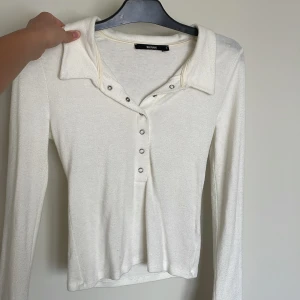 Vit långärmad topp med krage från BikBok - Säljer en stilren vit långärmad topp från BikBok med krage och knappar framtill. Toppen har en klassisk passform och är perfekt att matcha med jeans eller kjol.