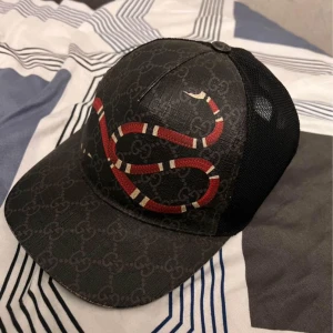 Svart Gucci keps med ormtryck - Snygg svart keps från Gucci med klassiskt GG-mönster och en stor röd och vit orm på framsidan. Baksidan är i mesh för extra ventilation och kepsen har justerbart spänne. Perfekt accessoar för en trendig look.
