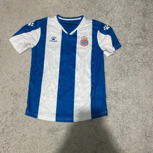 RCD Espanyol hemmatröja från Kelme - Säljer en officiell RCD Espanyol hemmatröja från Kelme. Tröjan är blå och vit med breda vertikala ränder, klubbmärke broderat på bröstet och Kelme-logga. Klassisk V-ringning och tillverkad i lätt, ventilerande material.