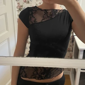 Festtopp med spetsdetaljer - Säljer en svart topp med spets. Toppen har korta ärmar och en asymmetrisk design vid halsen. Perfekt för dig som vill ha något unikt och trendigt till fester och sommaren
