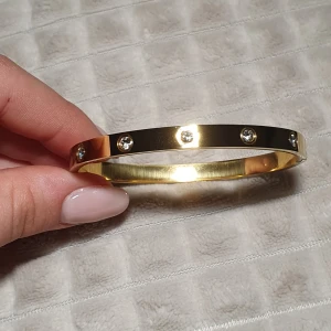 Guldfärgat armband med stenar - Snyggt och stilrent armband i guldfärg med infattade klara stenar runt om. Armbandet har en slät, polerad yta och är stelt i modellen. Perfekt för dig som gillar en elegant och tidlös accessoar. Armbandet är gjort i stainless steel.