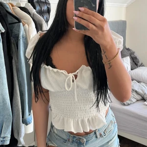 Vit croppad blus med volang och knyt - Säljer en söt vit croppad blus med volangkant och knyt framtill. Toppen har korta puffärmar och smockad midja som ger en feminin siluett. Perfekt till jeans eller kjol för en fräsch look.