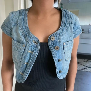 Jeans bolero  - Säljer en ljusblå croppad jeanbolero med korta ärmar och stora knappar framtill. Jackan har två bröstfickor och en snygg, öppen design. Perfekt för att lyfta en enkel outfit med lite edge.