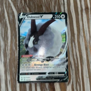 Pokémonkort Dubwool V - Säljer ett Pokémonkort med Dubwool V, 210 HP. Kortet har en holografisk yta och visar Dubwool mot en färgglad bakgrund. Kortet har attacker som Soft Wool och Revenge Blast. Perfekt för samlare eller Pokémonfans!